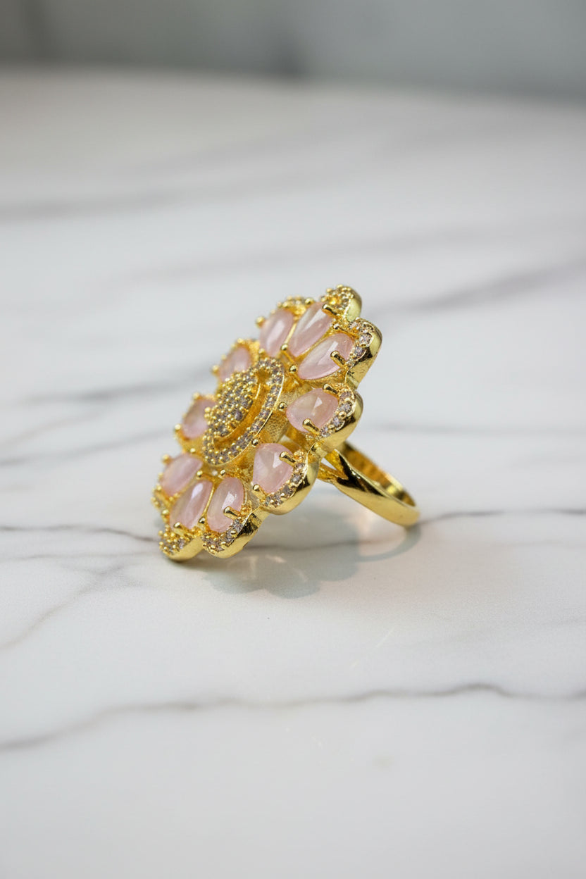 A gold-tone flower-shaped finger ring brooch with pale pink teardrop-shaped stones and a sparkling crystal centre, displayed on a white faux fur surface next to a brown textured bag.