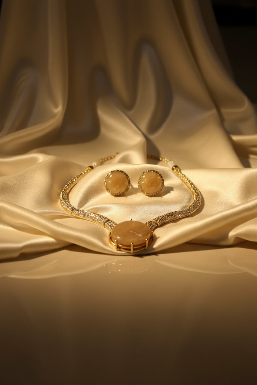 A gold-tone jewellery set with a matching necklace and earrings, featuring large oval beige cabochon stones and pavé-set clear rhinestones, held on a white card by a hand.