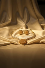 A gold-tone jewellery set with a matching necklace and earrings, featuring large oval beige cabochon stones and pavé-set clear rhinestones, held on a white card by a hand.