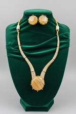 A gold-tone necklace and matching stud earrings displayed on a black velvet jewellery bust. The necklace features a large oval wooden pendant, textured round gold-toned studs, and small white bead accents.