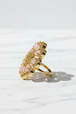A gold-tone ring with a floral cluster design featuring pale pink teardrop-shaped stones and small clear rhinestone accents, resting on a white faux fur surface.