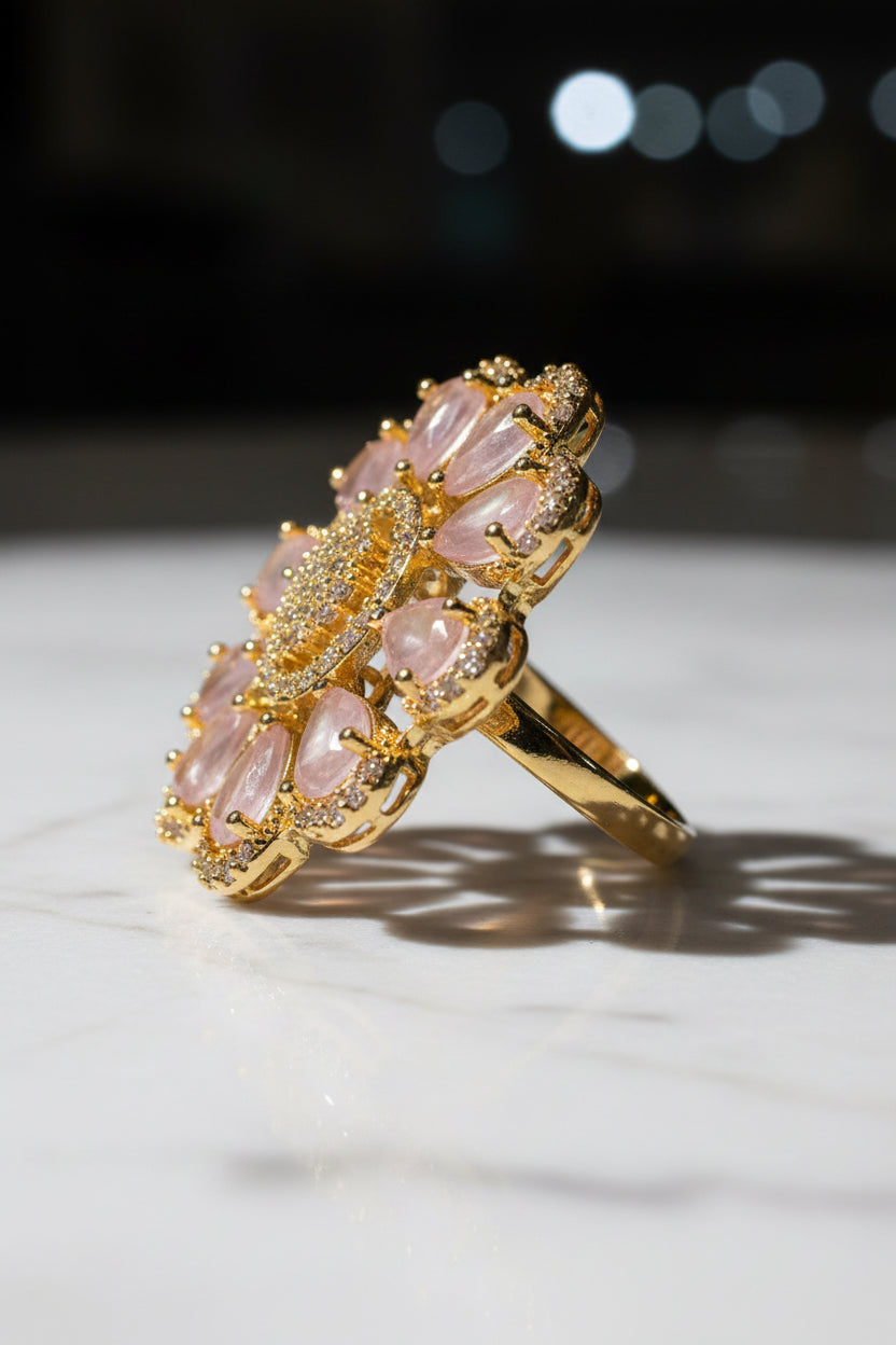 A gold-toned flower-shaped finger ring brooch with pale pink teardrop stones and a central cluster of clear stones, held in a hand against a soft white textured background.
