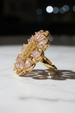 A gold-toned flower-shaped finger ring brooch with pale pink teardrop stones and a central cluster of clear stones, held in a hand against a soft white textured background.