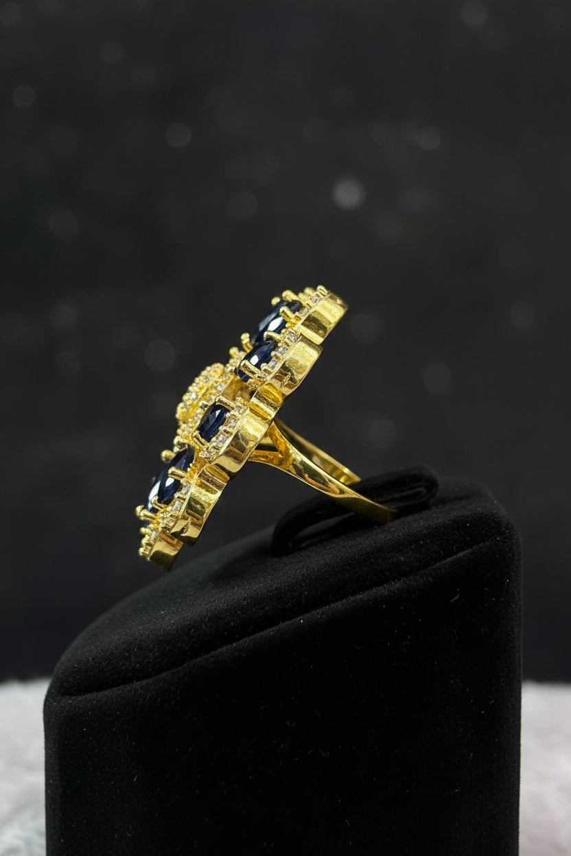 A gold-toned ring featuring multiple teardrop-shaped dark blue gemstones and small clear accent stones, displayed on a textured white faux fur surface.