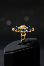 A gold flower-shaped brooch with deep blue teardrop gemstones and a central cluster of clear stones, resting on a white faux fur surface.