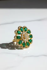 A gold flower-shaped finger ring brooch with green teardrop gemstones and a centre of clear stones, held by a hand against a white textured background.