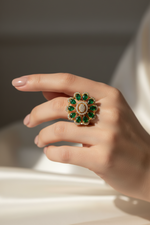 A close-up of a hand wearing a large floral ring with green emerald-cut stones and a gold-toned setting with small clear stones, photographed against a soft neutral background.