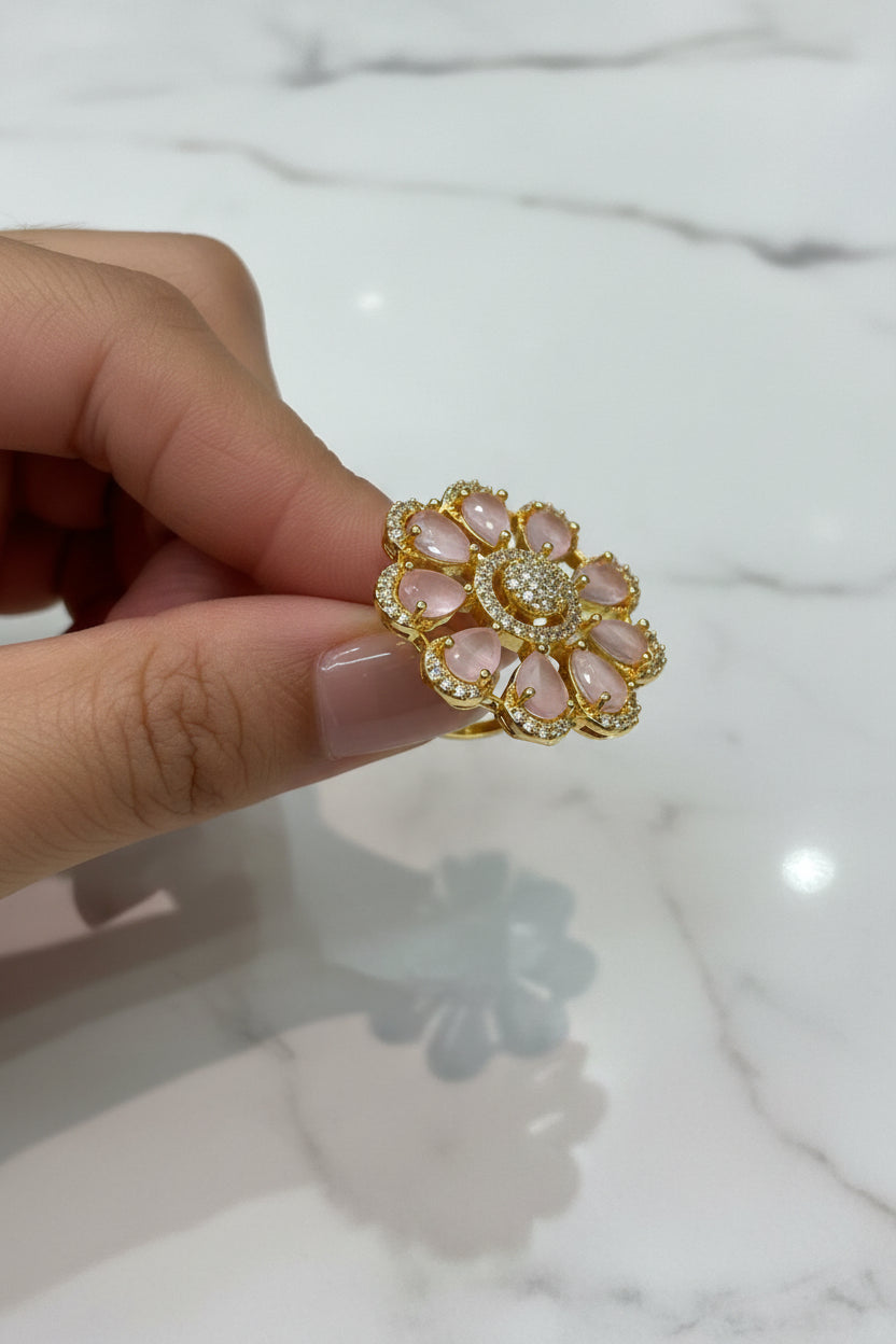 A gold flower-shaped finger ring brooch with pale pink teardrop stones and a central cluster of clear stones, held by a hand over a white textured surface.