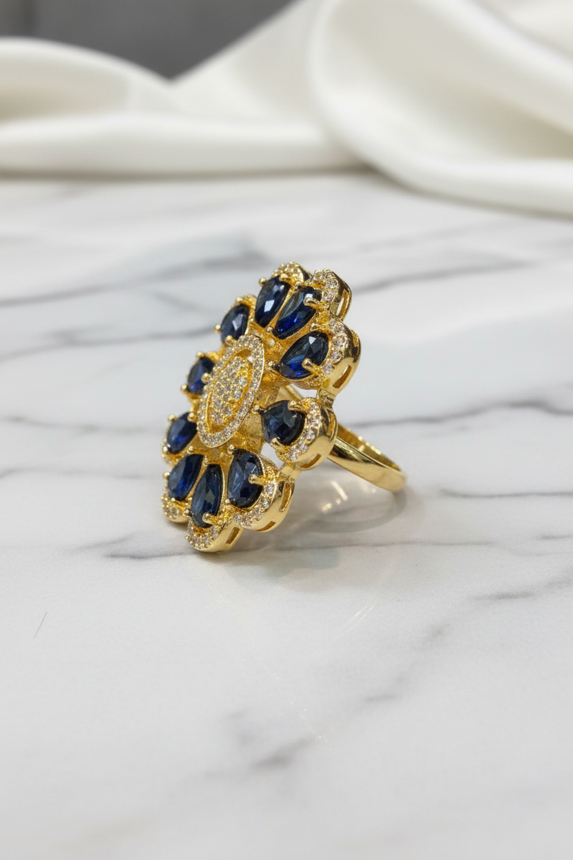 A gold ring with a floral design featuring deep blue gemstones and small clear stones, displayed on a white marble surface with a draped white fabric background.