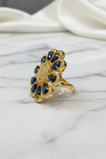 A gold ring with a floral design featuring deep blue gemstones and small clear stones, displayed on a white marble surface with a draped white fabric background.