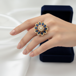 A gold flower-shaped ring with blue gemstones and a central clear stone, worn on a hand. A dark blue velvet ring box is visible in the background against a white draped fabric.