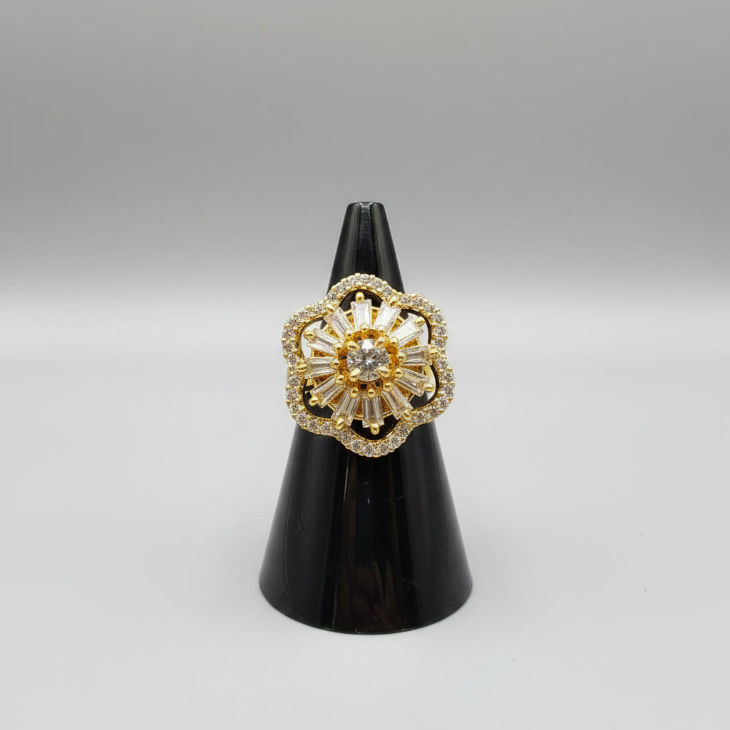 A gold flower-shaped ring with a large clear central gemstone and surrounding baguette and round stones, displayed on a black ring stand with a finger holding it.