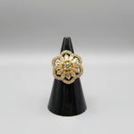 A gold flower-shaped ring with a large clear central gemstone and surrounding baguette and round stones, displayed on a black ring stand with a finger holding it.