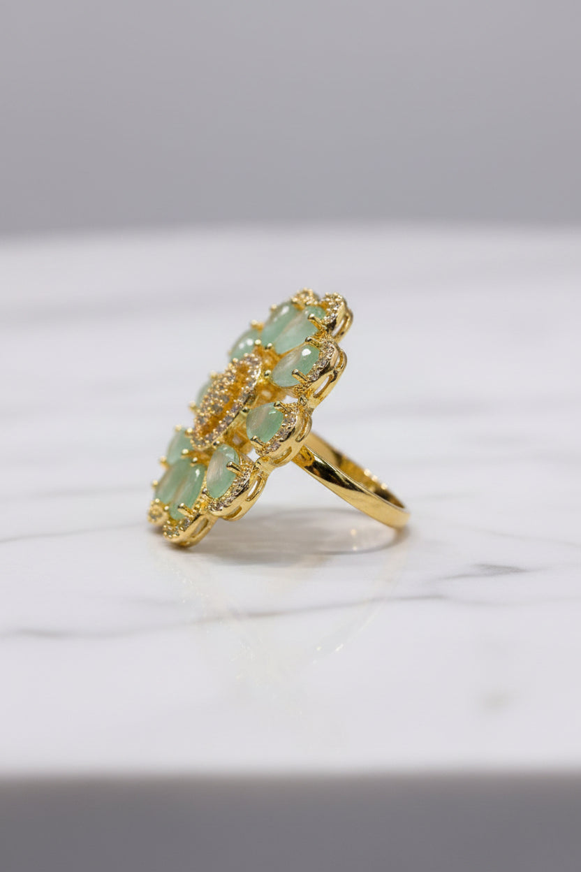 A gold flower-shaped ring with light green teardrop gemstones and a central cluster of small clear stones, held between fingers.