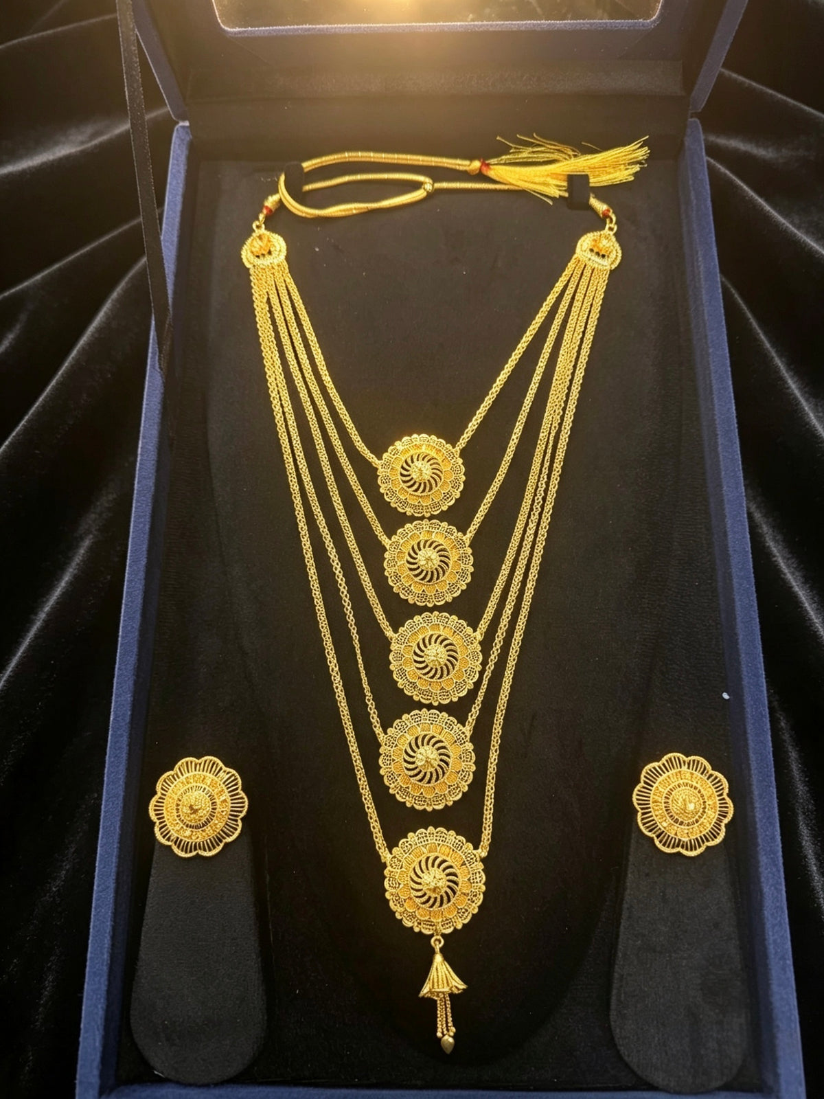 A gold jewellery set displayed inside a dark blue presentation box, featuring a multi-strand necklace with circular filigree motifs, a matching pendant, and two matching earrings on a black velvet insert.