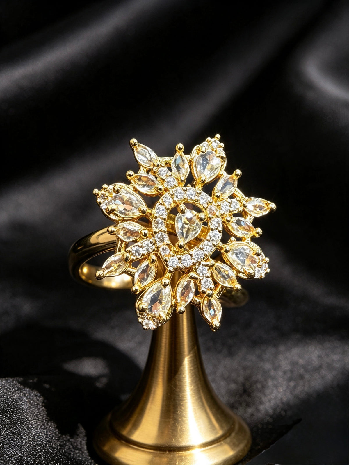 A gold ring with a large, ornate floral cluster of clear marquise and round stones, displayed on a gold stand against a black satin background.