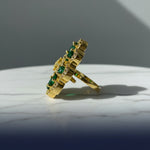 A gold ring with green gemstone accents, photographed on a textured gray faux fur surface.