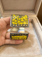 A hand holding a wide cuff bracelet with a yellow and black mosaic tile pattern and gold-tone studs, positioned in front of an open beige jewellery box.