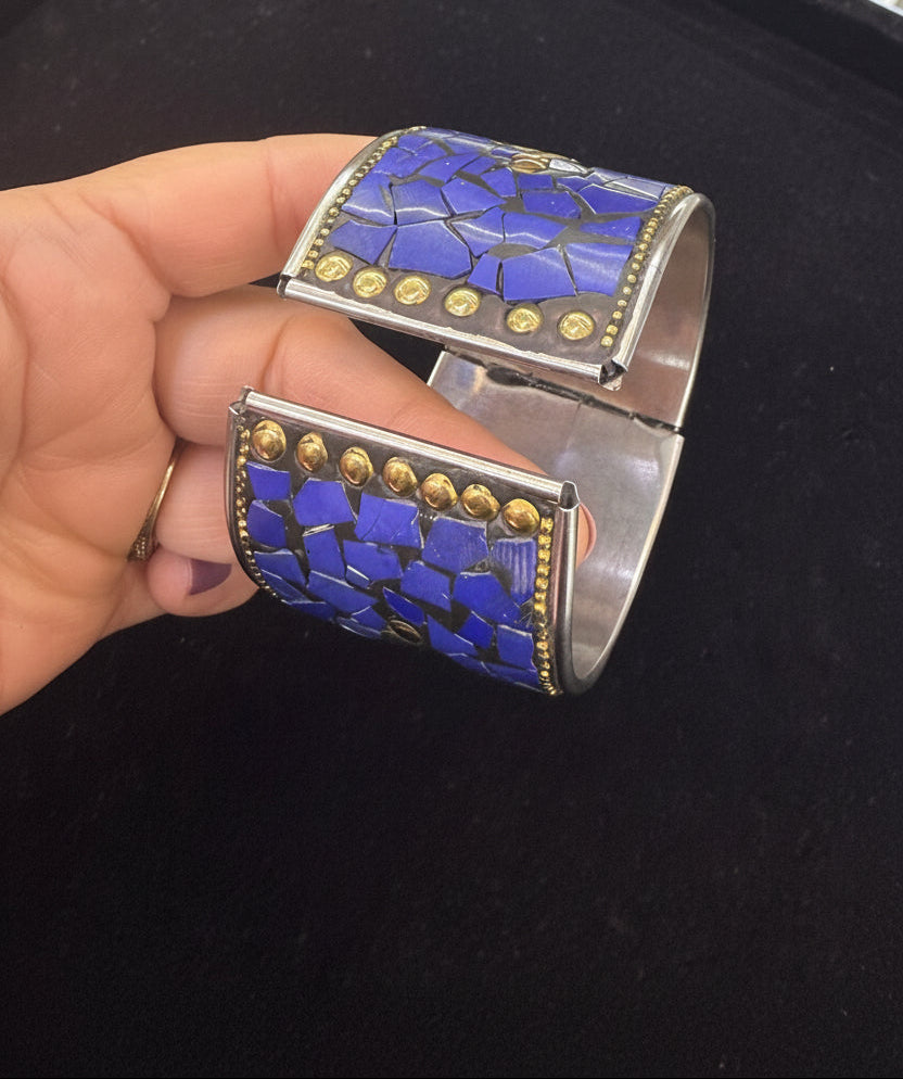 A hand holding a wide silver cuff bracelet with a blue mosaic stone inlay and gold-tone studs and border detailing, shown against a dark background.