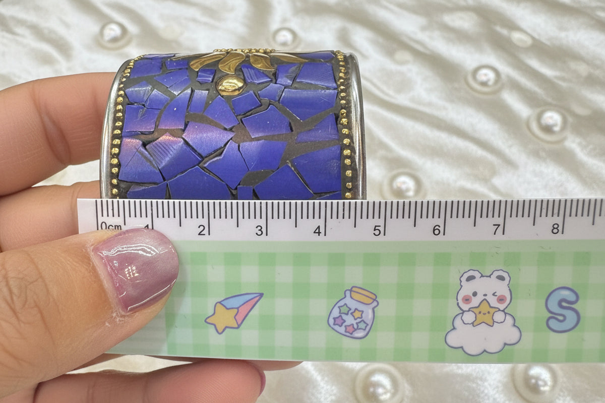 A hand holds a decorative blue and gold metallic cuff bracelet next to a green measuring tape for scale, with a jewellery display case in the background.