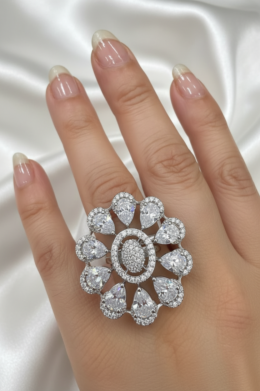 A close-up of a hand wearing a large floral ring made of clear gemstones in a silver-toned setting, against a black background.