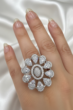 A close-up of a hand wearing a large floral ring made of clear gemstones in a silver-toned setting, against a black background.