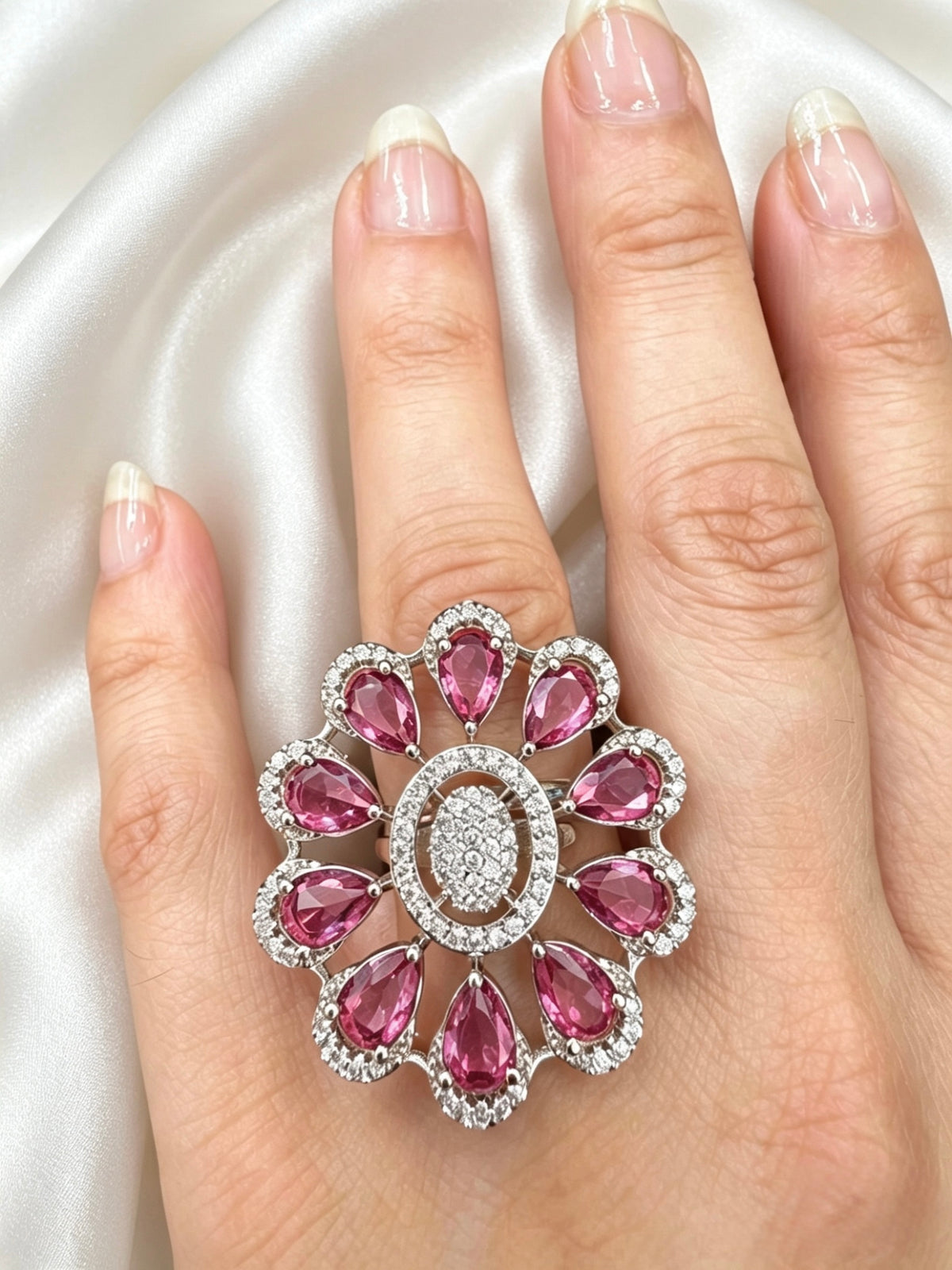 A large floral statement ring with pink teardrop gemstones and a central oval cluster of clear stones, shown worn on a hand against a satin fabric background.