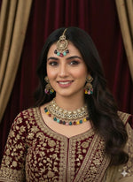 A model in a maroon embroidered velvet outfit wearing a luxury kundan jewelry set with vibrant, multi-colored gemstone drops against a dark velvet backdrop.