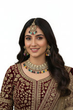 A model in a maroon embroidered velvet outfit wearing a luxury kundan jewelry set with vibrant, multi-colored gemstone drops against a dark velvet backdrop.