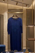 A navy blue embroidered dress displayed on a hanger inside a luxury boutique, with jewellery and a folded matching blue scarf on a marble counter below.