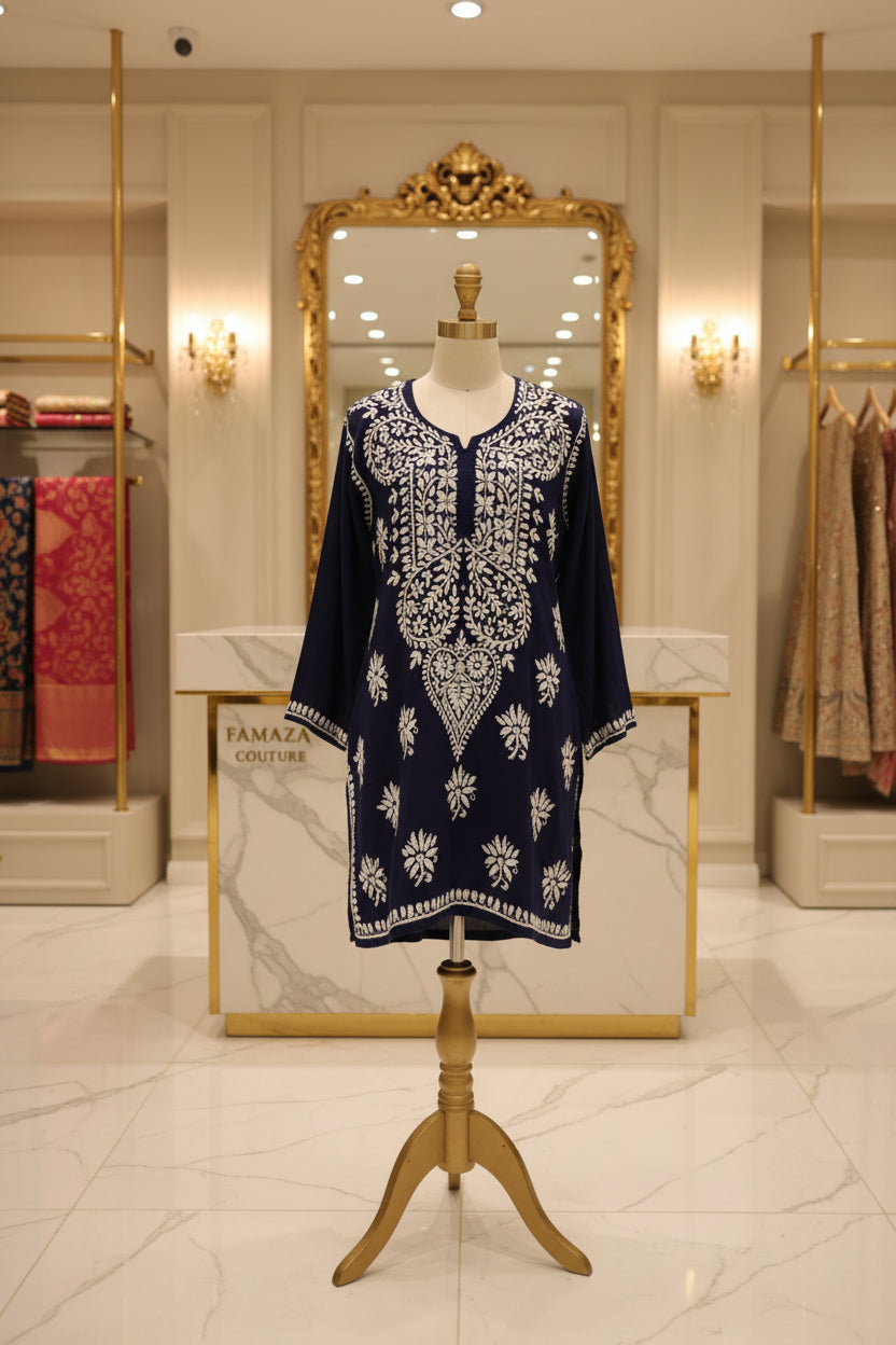 A navy blue long-sleeve short tunic with intricate white embroidery, laid flat on a grey herringbone wood floor in a retail store.