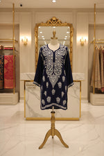 A navy blue long-sleeve short tunic with intricate white embroidery, laid flat on a grey herringbone wood floor in a retail store.