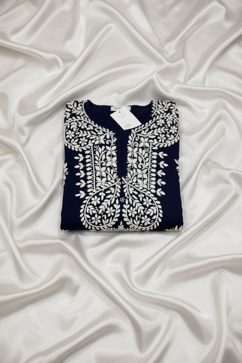 A navy blue tunic top with intricate white embroidery and floral motifs, laid flat on a grey wood floor with a white tag still attached at the collar.