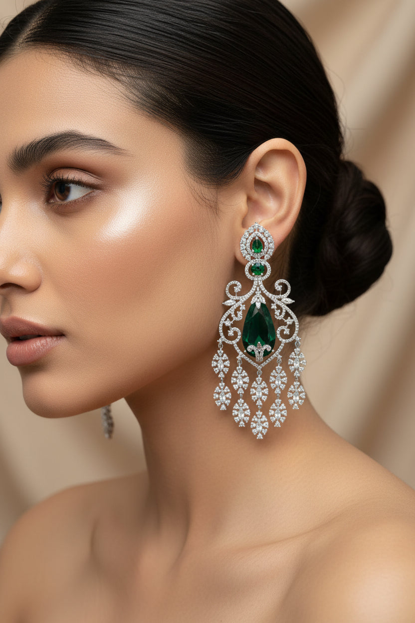 A pair of American diamond ornate silver-toned chandelier earrings with large green teardrop-shaped center stones and smaller green stones above, embellished with clear rhinestones, displayed on a light wood surface.