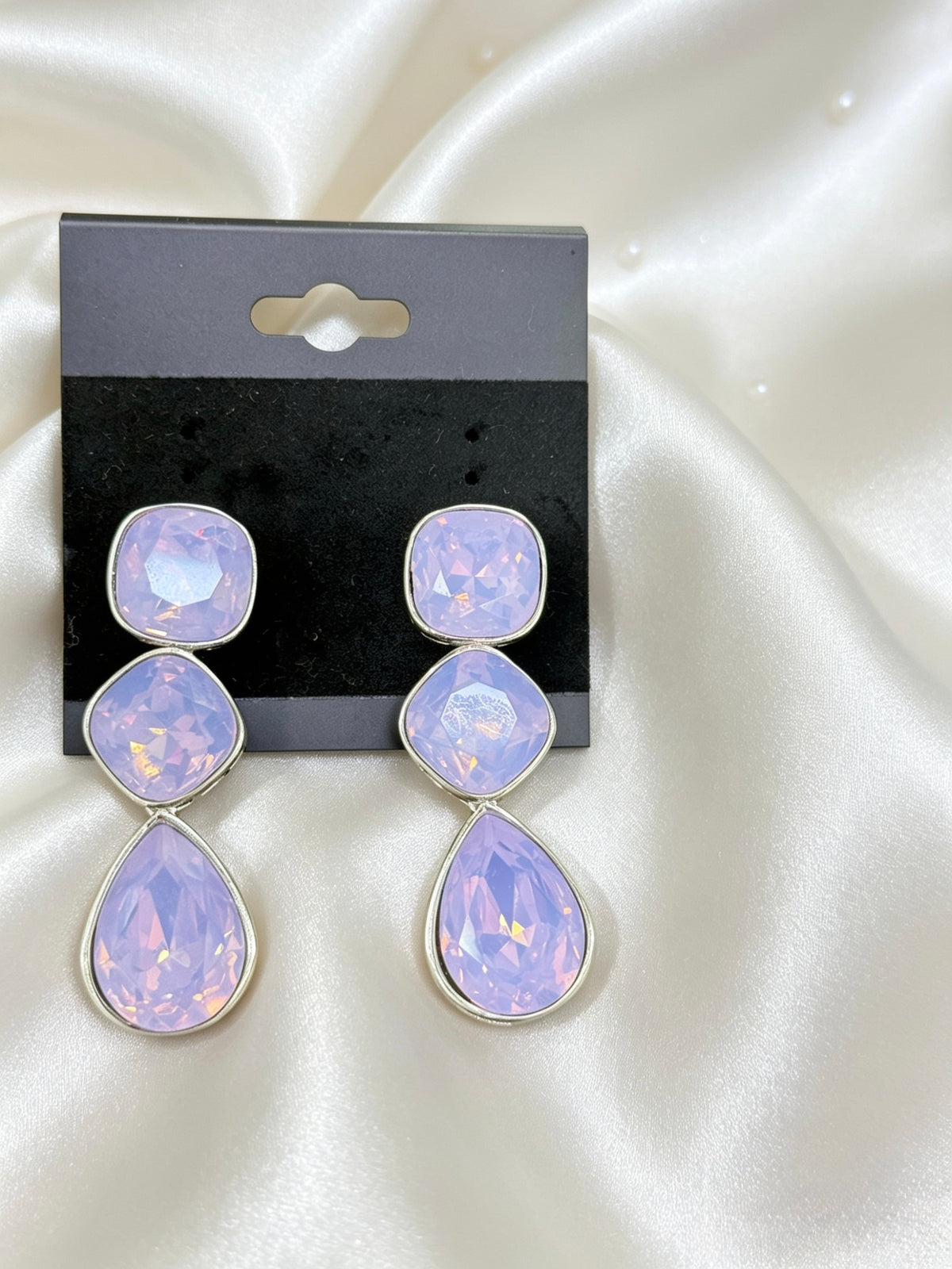 A pair of dangling earrings with three stacked oval-cut light purple stones in silver settings, displayed on a black retail card against a white satin fabric background.