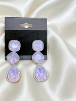 A pair of dangling earrings with three stacked oval-cut light purple stones in silver settings, displayed on a black retail card against a white satin fabric background.