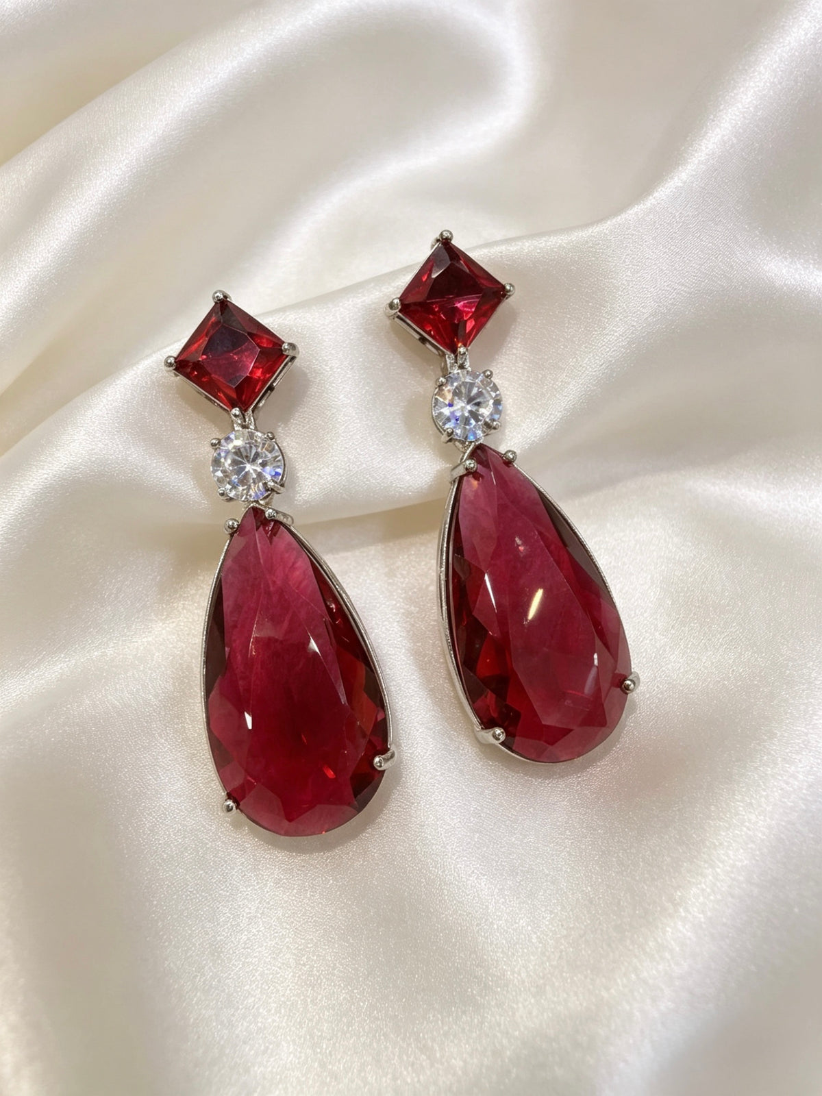 A pair of drop earrings with deep red teardrop-shaped gemstones, square red stones at the top, and small clear round stones in between, displayed on a draped cream satin fabric.