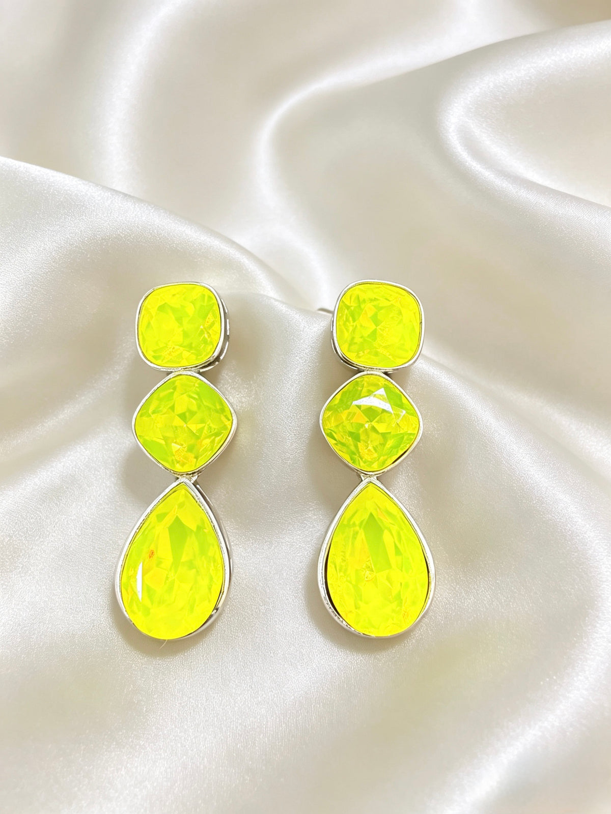 A pair of drop earrings with three stacked bright neon yellow gemstones in silver settings, displayed on a draped white satin fabric background.