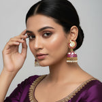 A pair of gold-plated Meenakari peacock Jhumka earrings with purple and white enamel detailing, displayed on a black stand.