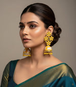 A pair of gold-plated brass jhumka earrings with yellow stones and gold meenakari work displayed on a stand.