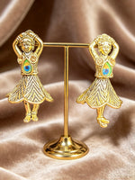 A pair of gold-toned earrings shaped like dancing girls, displayed on a small gold stand against a draped satin fabric background.
