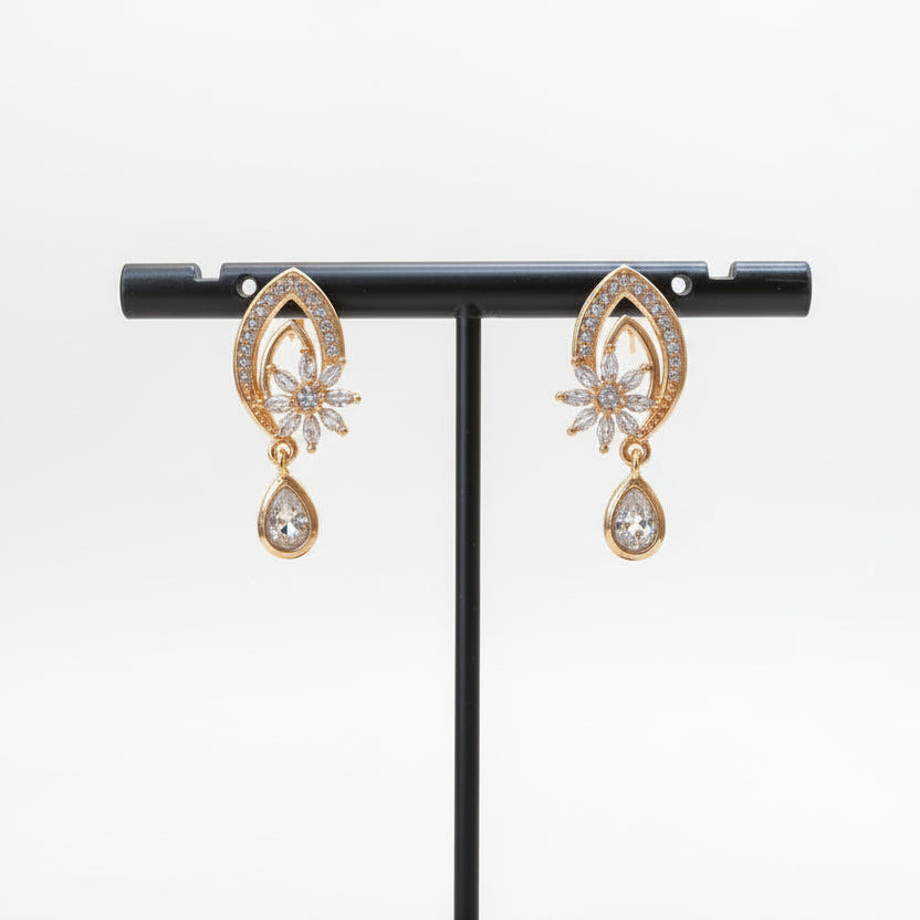 A pair of golden brass earrings featuring an intricate, petal-shaped frame encrusted with small crystals and a dangling, clear teardrop charm.