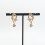A pair of golden brass earrings featuring an intricate, petal-shaped frame encrusted with small crystals and a dangling, clear teardrop charm.