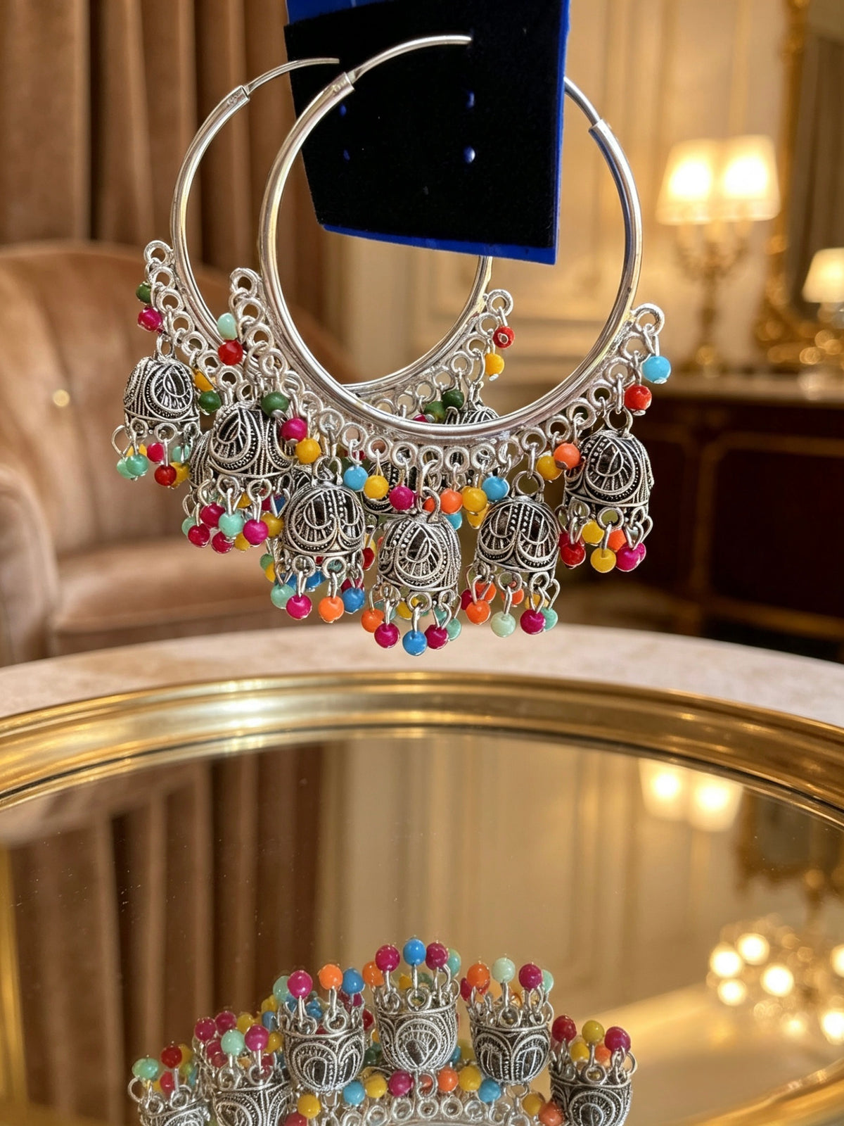 A pair of large silver-tone hoop earrings with ornate filigree dangles and colorful bead accents, shown hanging above a mirrored surface in an indoor setting.