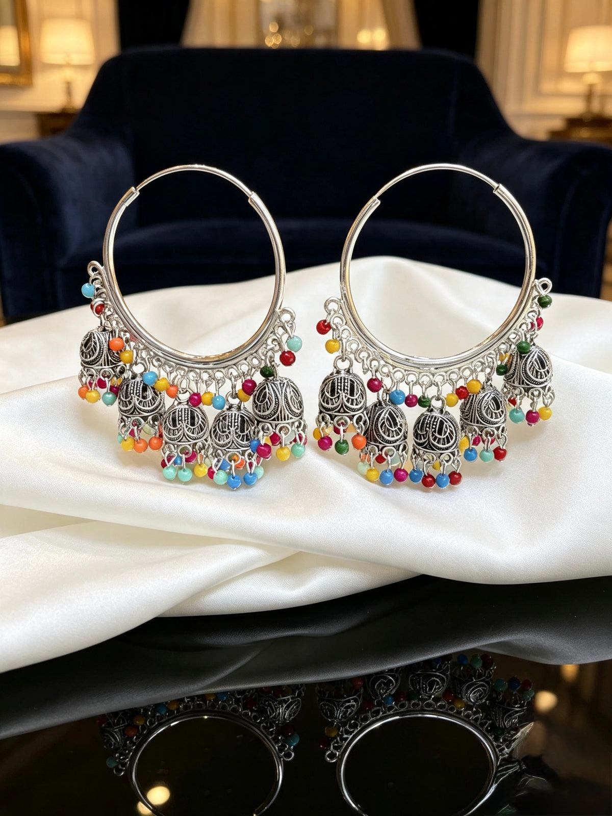 A pair of large silver-toned hoop earrings with ornate dangling filigree and many small multi-coloured bead accents, displayed on draped white satin fabric with a dark blue upholstered chair in the background.