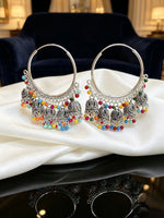A pair of large silver-toned hoop earrings with ornate dangling filigree and many small multi-coloured bead accents, displayed on draped white satin fabric with a dark blue upholstered chair in the background.