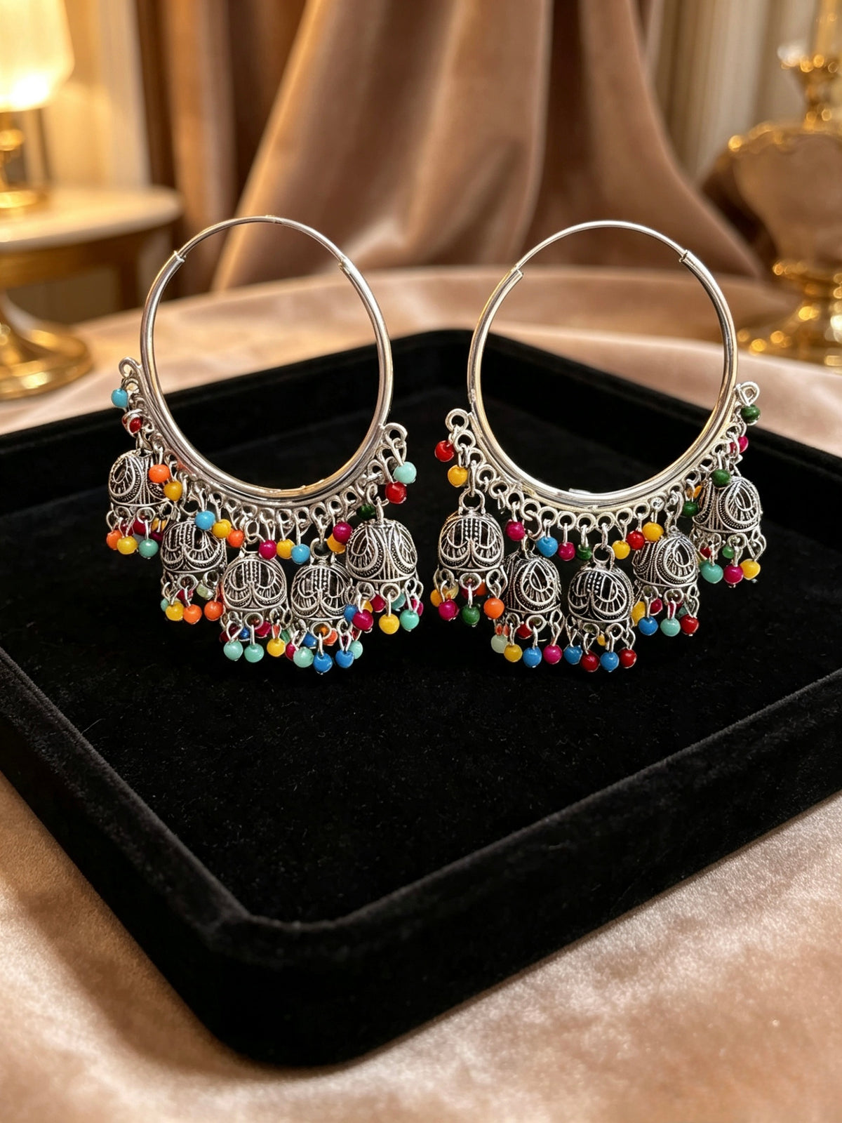 A pair of large silver-toned hoop earrings with ornate dangling filigree charms and small multi-coloured beads, displayed on a black velvet jewellery tray.