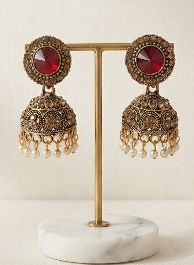 A pair of ornate Indian jhumka earrings with red circular stones and gold-toned filigree bells with pearl-like dangling beads, displayed on a gold stand with a white marble base.