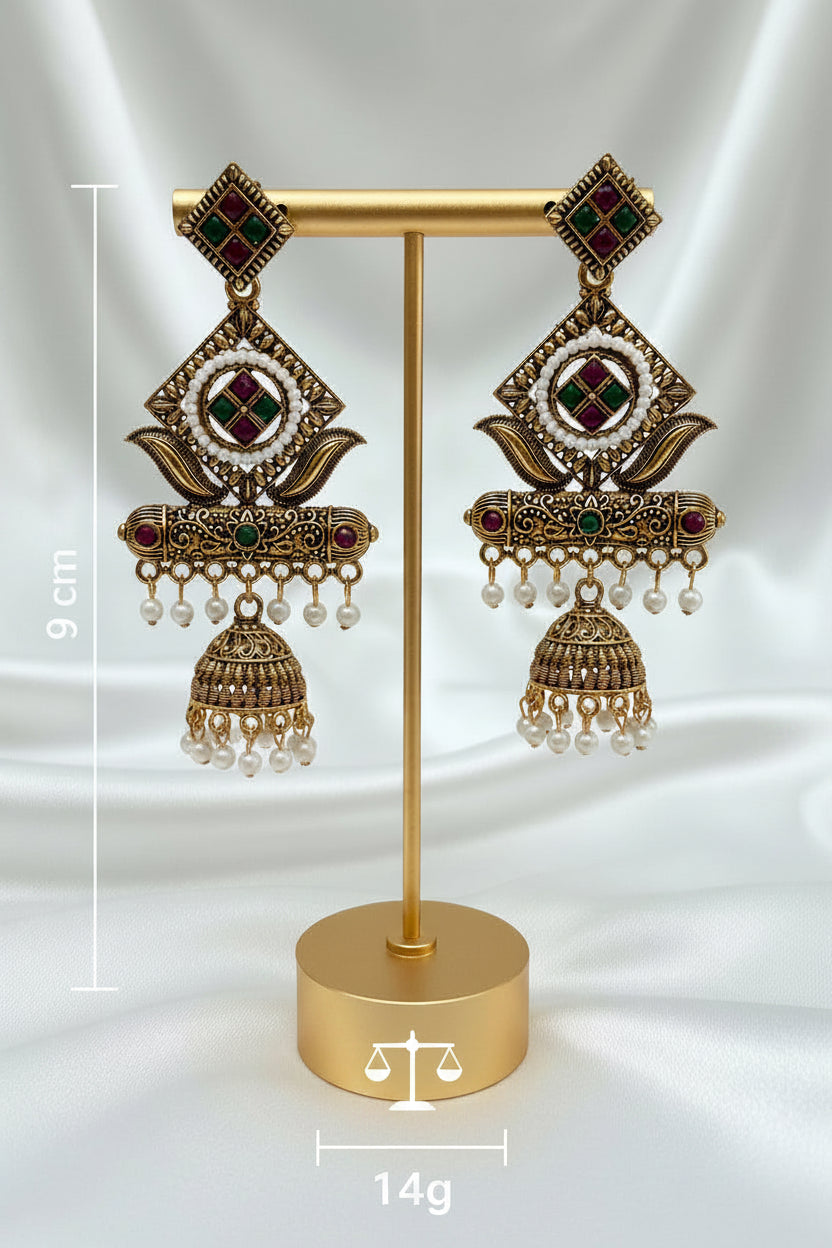 A pair of ornate gold-toned Indian jhumka earrings with white pearl bead borders, green and red enamel details, and dangling pearl beads, displayed on a light marble surface.