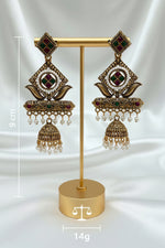 A pair of ornate gold-toned Indian jhumka earrings with white pearl bead borders, green and red enamel details, and dangling pearl beads, displayed on a light marble surface.